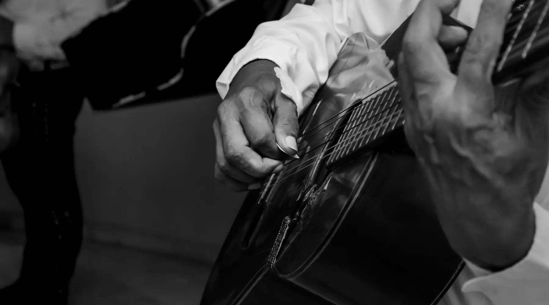 Ruce flamenkového kytaristy na černé kytaře, černobíle, vedle něj tmavé kalhoty dalšího hudebníka.
