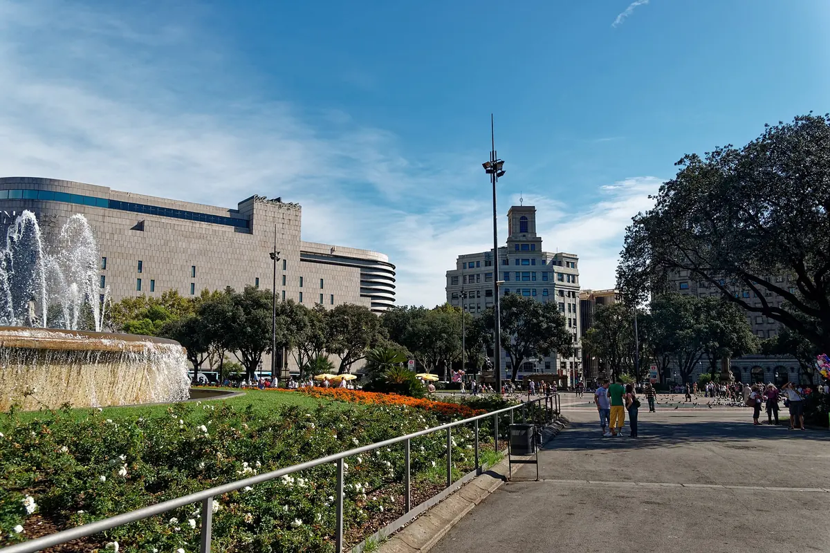 Fontána a květinové záhony na Plaça de Catalunya v Barceloně, v pozadí věž a stromy.