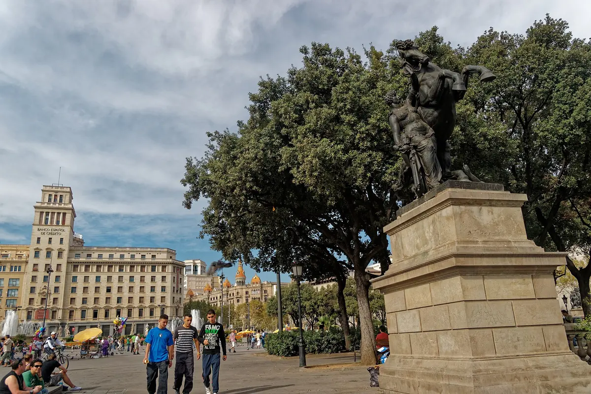 Bronzová socha na kamenném podstavci pod stromy na Plaça de Catalunya, kolem procházejí lidé a v pozadí stojí kamenná věž.