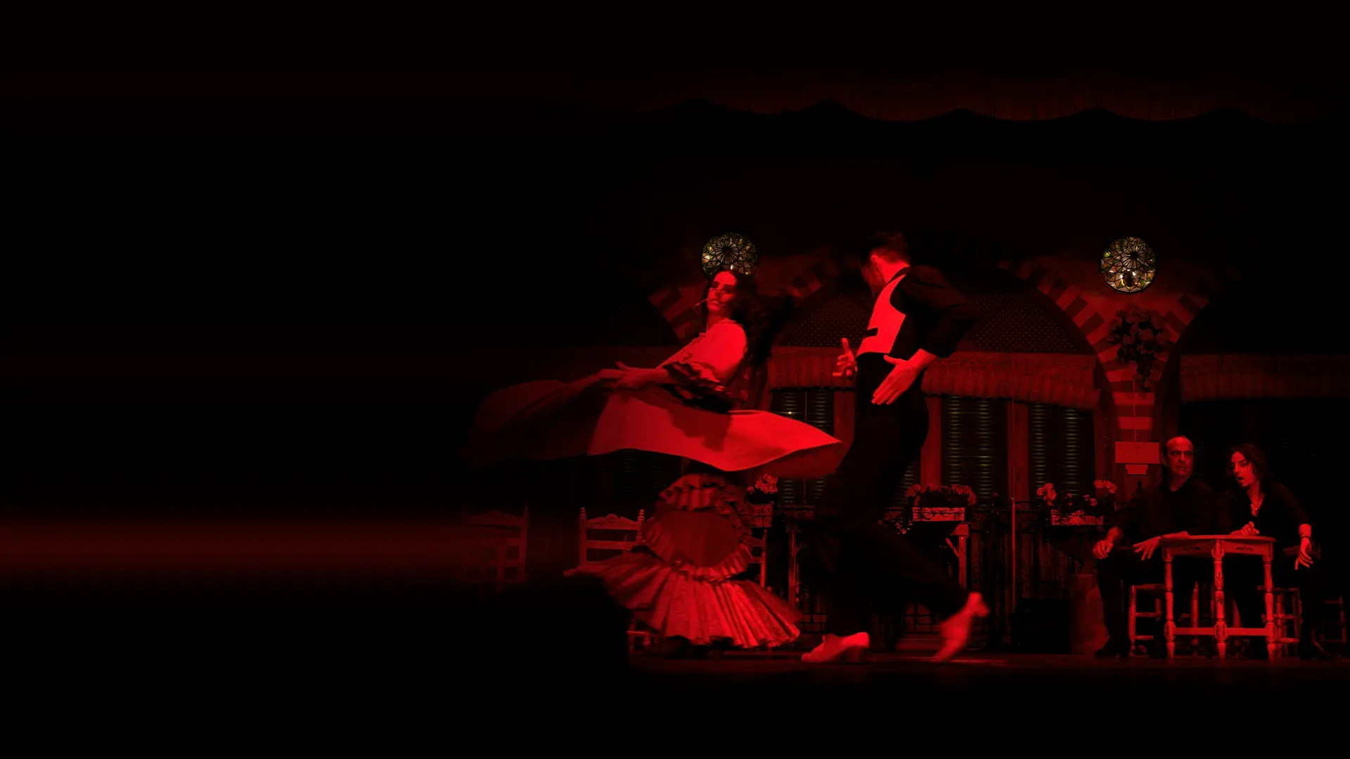 Flamenková tanečnice v červené sukni se otáčí vedle tanečníka v černé vestě na jevišti s klenutou andaluskou kulisou, vpravo sedí dva hudebníci.